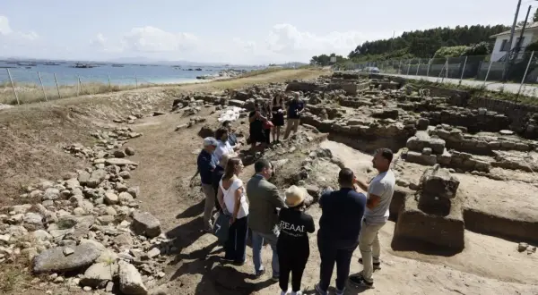 Grupo de personas observando ruinas arqueológicas en la costa. Se aprecia el mar y el **adro vello** del yacimiento.