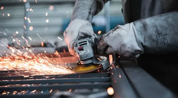 Pracownik w rękawicach szlifuje metal, sypią się iskry. Uważaj na wypadek przy pracy!
