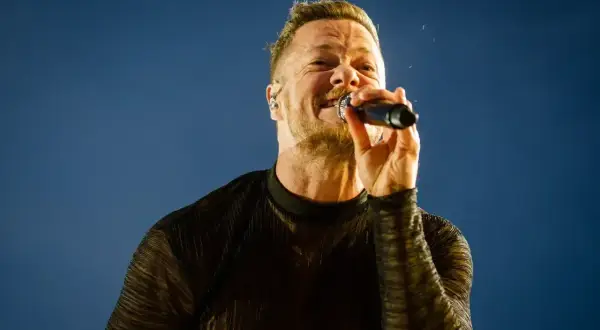 Wokalista Imagine Dragons śpiewa do mikrofonu na tle błękitnego nieba.