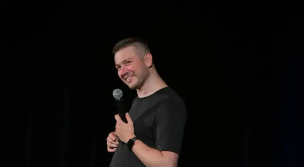 Grzegorz Wójtowicz w trakcie występu stand-up, trzyma mikrofon i uśmiecha się do publiczności.