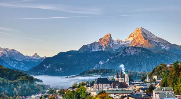 Morgensonne beleuchtet den spitzen Gipfel des kleinen Watzmanns über einem nebelverhangenen Tal und einer malerischen Stadt mit Kirchtürmen.