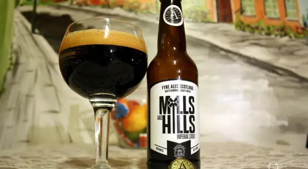 Szklanka z ciemnym imperial stout i butelka piwa Mills and Hills na tle malarskiej sceny.
