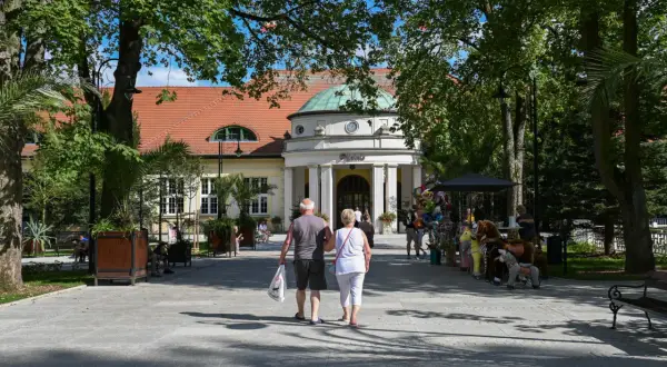 Para starszych osób spaceruje alejką przed budynkiem sanatorium.
