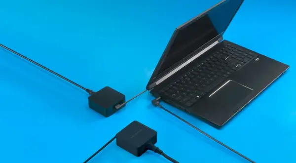 Czarny laptop gamingowy podłączony do dwóch zasilaczy na niebieskim tle.