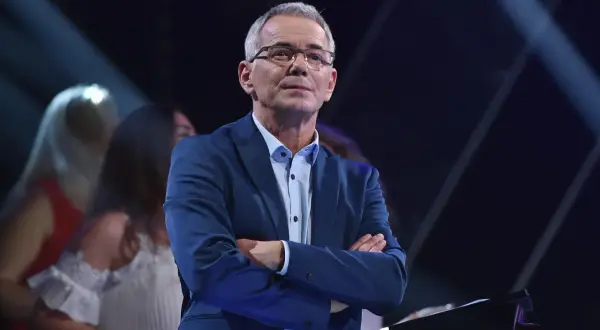 Robert Janowski w garniturze, z rękami skrzyżowanymi na piersi, na tle sceny. Czy to właśnie powód, dlaczego Robert Janowski naprawdę odszedł z Jaka to melodia po latach?