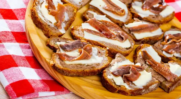 Kromki chleba z masłem i filecikami anchois ułożone na drewnianej desce, obok czerwono-biała serwetka.
