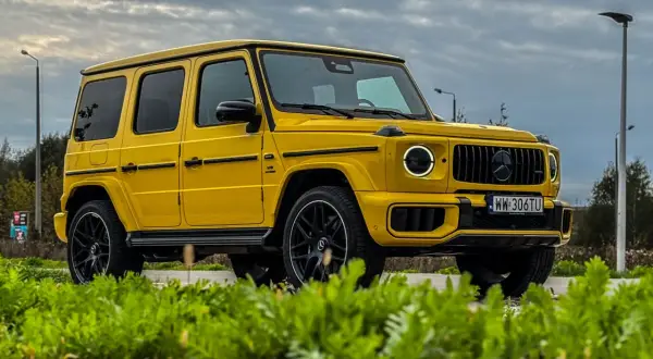 Żółty Mercedes-Benz G63 AMG z czarnymi felgami i przyciemnianymi szybami, zaparkowany na tle pochmurnego nieba i zieleni.