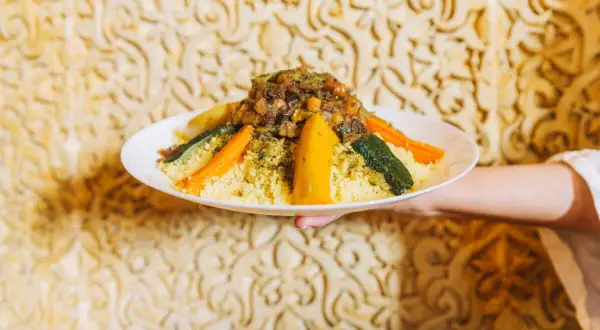 Un plat de couscous algérien, garni de légumes colorés et d'une viande savoureuse, est présenté sur un fond orné.