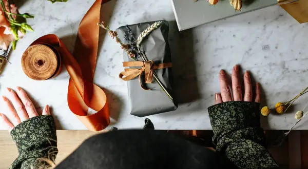 Händer som förbereder sig för att slå in runda paket med grått papper, orange sidenband och torkade blommor.