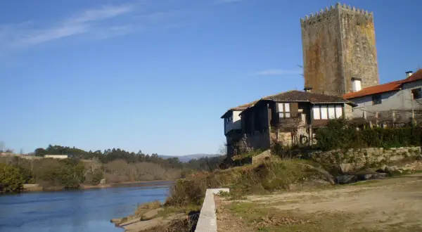 La torre de Lapela se alza junto a un río, con casas antiguas a su lado y un cielo azul despejado.