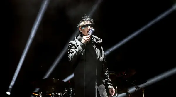 Marilyn Manson na scenie w Warszawie, w czarnym płaszczu i z charakterystycznym makijażem, śpiewa do mikrofonu.