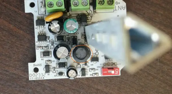 Uszkodzona lampa ostrzegawcza bramy. Widoczny element elektroniczny z okrągłym, czarnym uszkodzeniem.
