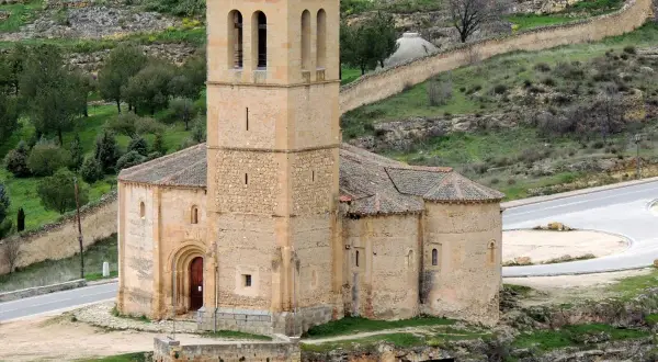 La iglesia de la Veracruz, de piedra y con un campanario robusto, se alza sobre un paisaje verde y ondulado, con un camino serpenteante a su lado.