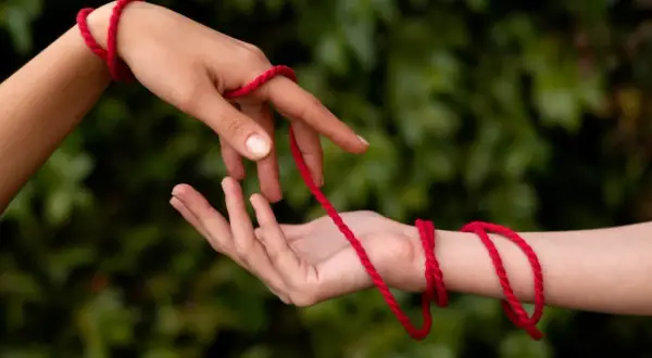 Dwie dłonie splatają czerwoną nitkę. Czy czerwona nitka to grzech? Symbol połączenia, losu, a może czegoś więcej?