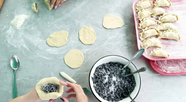 Ręce formują ciasto wonton z jagodami i cukrem. Na blacie leżą okrągłe placki ciasta, miska z jagodami i tacka z gotowymi pierożkami.