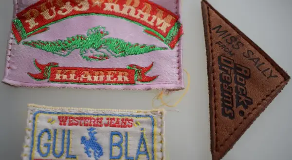 Tre märken: ett rosa med texten "PUSS & KRAM JEANS" och en grön drake, ett brunt triangelformat med "MISS SALLY FROM Rock Dreams", och ett vitt med "WESTERN JEANS GUL BLÅ WYOMING".