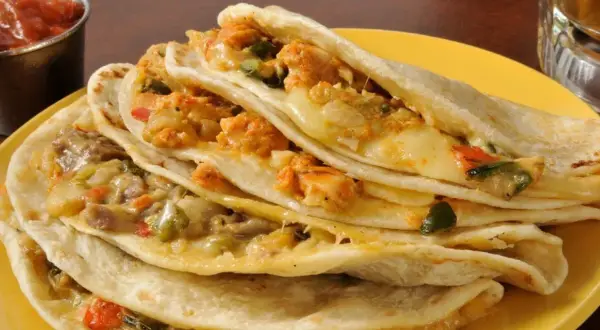 Quesadillas de pollo apiladas, rellenas de queso derretido y trozos de pollo sazonado, servidas en un plato amarillo con salsa al lado.