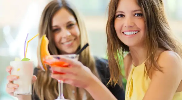 Dwie uśmiechnięte kobiety z drinkami. Jedna trzyma biały koktajl, druga pomarańczowy. Czy wódka tuczy?