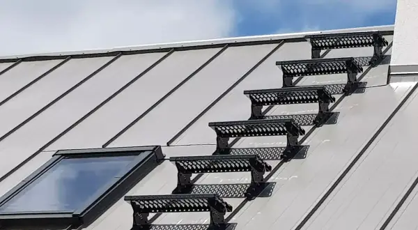 Sześć czarnych stopni bezpieczeństwa na metalowym dachu, prowadzących do okna dachowego. Solidne opierzenie dachu zapewnia bezpieczne przejście.