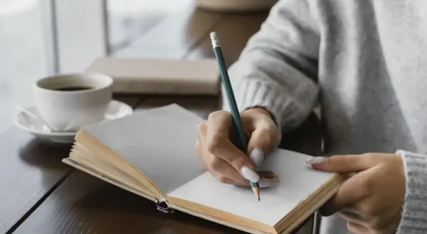 Mano escribiendo en un diario, un ritual de autocuidado. El café humeante acompaña este momento de introspección, una forma de escribir un diario como terapia.