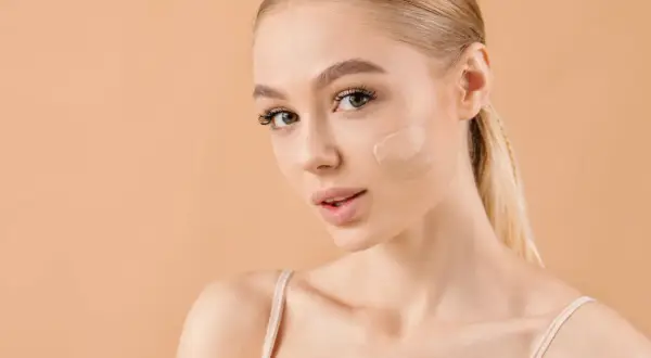 Kobieta z blond włosami pokazuje, gdzie nakładać korektor na policzek. Idealny odcień dla promiennej cery.