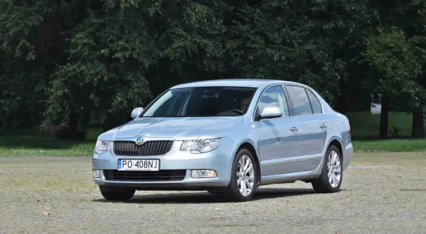 Jasnoniebieska Skoda Superb 2 z nowymi żarówkami, zaparkowana na placu przed drzewami.