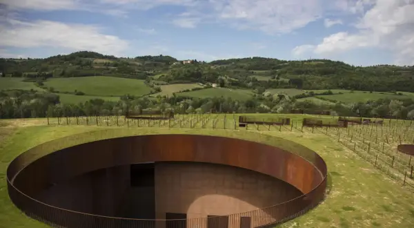 Un cerchio di metallo arrugginito emerge dal verde delle colline del Chianti, con vigneti che si estendono all'orizzonte sotto un cielo azzurro.