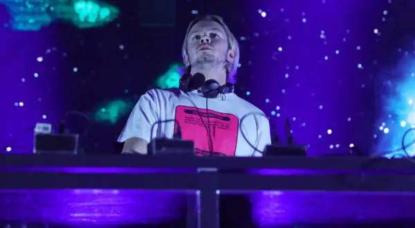 DJ na scenie, w słuchawkach na szyi, z napisem na koszulce. W tle niebieskie światła i efekty. To część lineupu Wisłoujście.