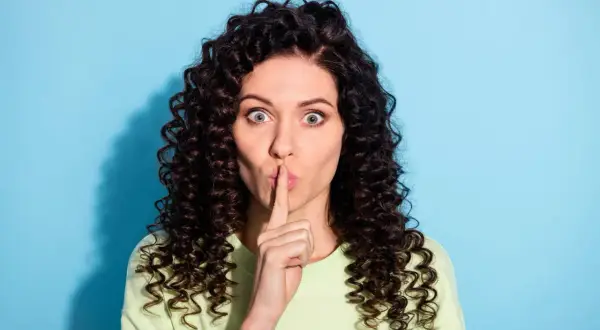Vrouw met krullend haar houdt vinger voor de mond. Ze zegt niets, want verzwijgen is liegen.