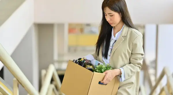 Mujer asiática con caja de pertenencias, gafas en el cuello, caminando por escaleras. Su expresión es de tristeza, quizás por un despido abreviado.