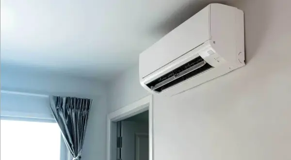 Biała jednostka klimatyzacyjna zamontowana na jasnej ścianie, obok okna z szarą zasłoną.
