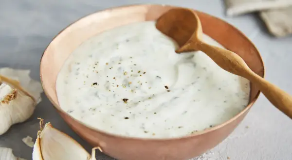 Ein cremiger Knoblauch Joghurt Dip in einer Kupferschale, garniert mit Pfeffer und Kräutern. Daneben liegen frische Knoblauchzehen.