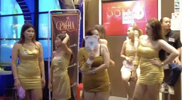 Kobiety w złotych sukienkach pozują przed banerem "Pacha". W tle neonowy napis "Shots". To scena z nocnego życia, być może związana z ping pong show w Tajlandii.