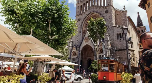 En charmig spårvagn passerar framför en kyrka i Soller, Palma. Folk sitter vid uteserveringar under parasoll.