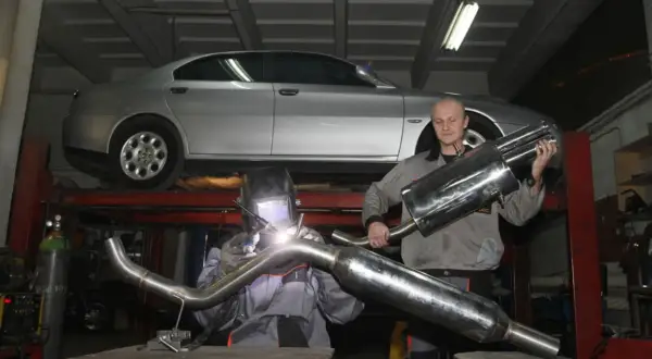 Mechanik w masce spawalniczej wykonuje spawanie wydechu. Obok stoi kolega z gotową częścią.