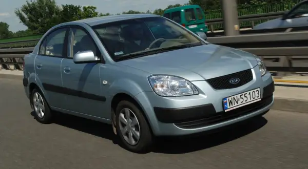 Błękitne światła Kia Rio na drodze. Kierowca w okularach prowadzi auto. W tle zielony van i drzewa.