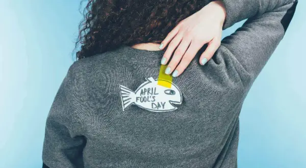 Ręka z pastelowym lakierem przyczepia do swetra papierową rybę z napisem "April Fool's Day".