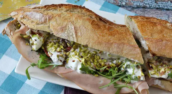 Un sandwich baguette garni de jambon, roquette, tomates séchées, pistaches concassées et burrata crémeuse.