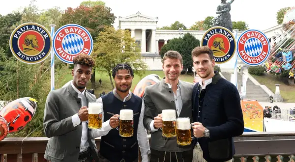 Vier FC Bayern Spieler in Tracht mit Bierkrügen, vor Paulaner- und FC Bayern-Logos. Ein Fest für alle Fans bei Veranstaltungen in Bayern.