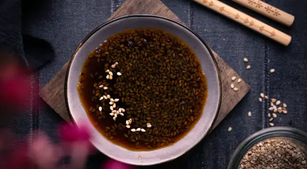 Schale mit Teriyaki-Sauce und Sesam, daneben Stäbchen und ein Glas Sesamsamen. Perfekt für dein Teriyaki Sauce Rezept!
