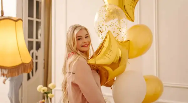 Uśmiechnięta blondynka z bukietem złotych i białych balonów w kształcie gwiazd i kuli.