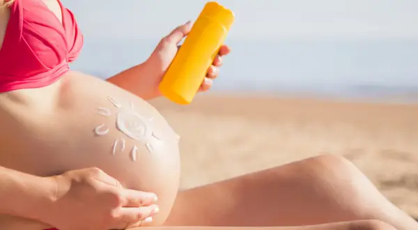 Mujer embarazada en la playa aplica protector solar en su barriga, dibujando un sol. Cuidando su piel durante el embarazo.