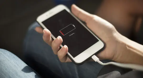 Dłoń trzyma smartfon z ikoną niskiego poziomu baterii i napisem "Charging". Wtyczka kabla ładującego jest podłączona. Oszczędzanie baterii jest kluczowe.