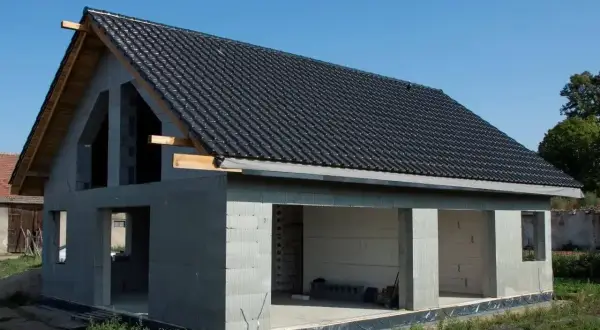 Dom w stanie surowym otwartym, z betonowych pustaków, z czarnym dachem i otwartymi otworami okiennymi i drzwiowymi.