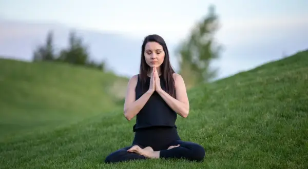 Mujer en posición de loto en la hierba, con las manos juntas en oración. Su rutina de yoga al aire libre es un momento de paz.