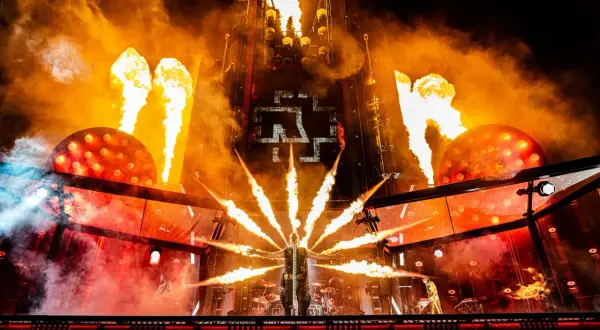 Płomienie buchają na koncercie Rammstein, tworząc piekielną atmosferę. Wokalista stoi w centrum, otoczony ogniem.