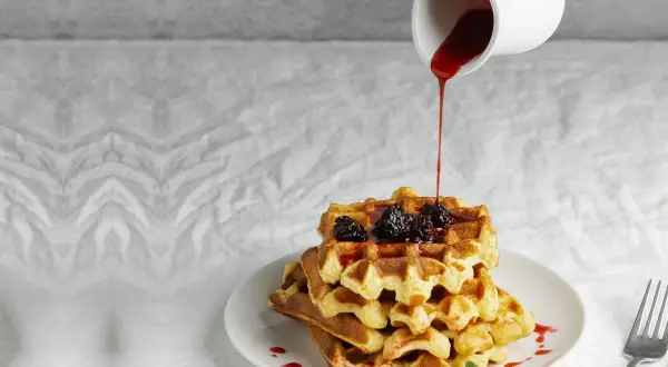 Pila di waffle dorati con more e sciroppo rosso versato da una caraffa. Ecco come fare i waffle perfetti per la colazione.