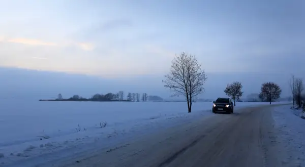 Samochód jedzie po ośnieżonej drodze wśród pól. Wczesny wieczór, niebo lekko zaróżowione. Jazda po śniegu wymaga ostrożności.