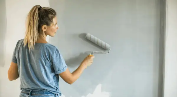 Donna con i capelli biondi in coda di cavallo, jeans e maglietta blu, impara come pitturare con il rullo una parete grigia.