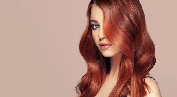 Ein Porträt einer Frau mit voluminösen, dunkelroten Haaren, die ihr Gesicht umspielen. Ihre blauen Augen blicken nachdenklich in die Ferne.